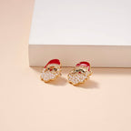 The Pearl Santa Claus Stud Earrings by Lilla Haven, featuring gold studs with red hats and white beaded beards, rest on a cream surface beside a white rectangular object—a charming Christmas holiday accessory and the perfect festive gift.