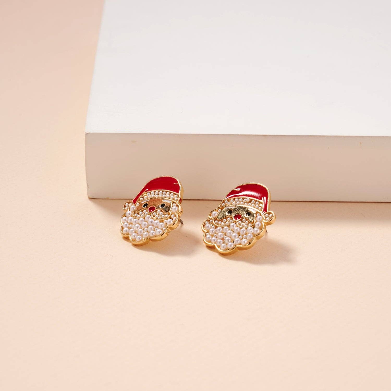 The Pearl Santa Claus Stud Earrings by Lilla Haven, featuring gold studs with red hats and white beaded beards, rest on a cream surface beside a white rectangular object—a charming Christmas holiday accessory and the perfect festive gift.