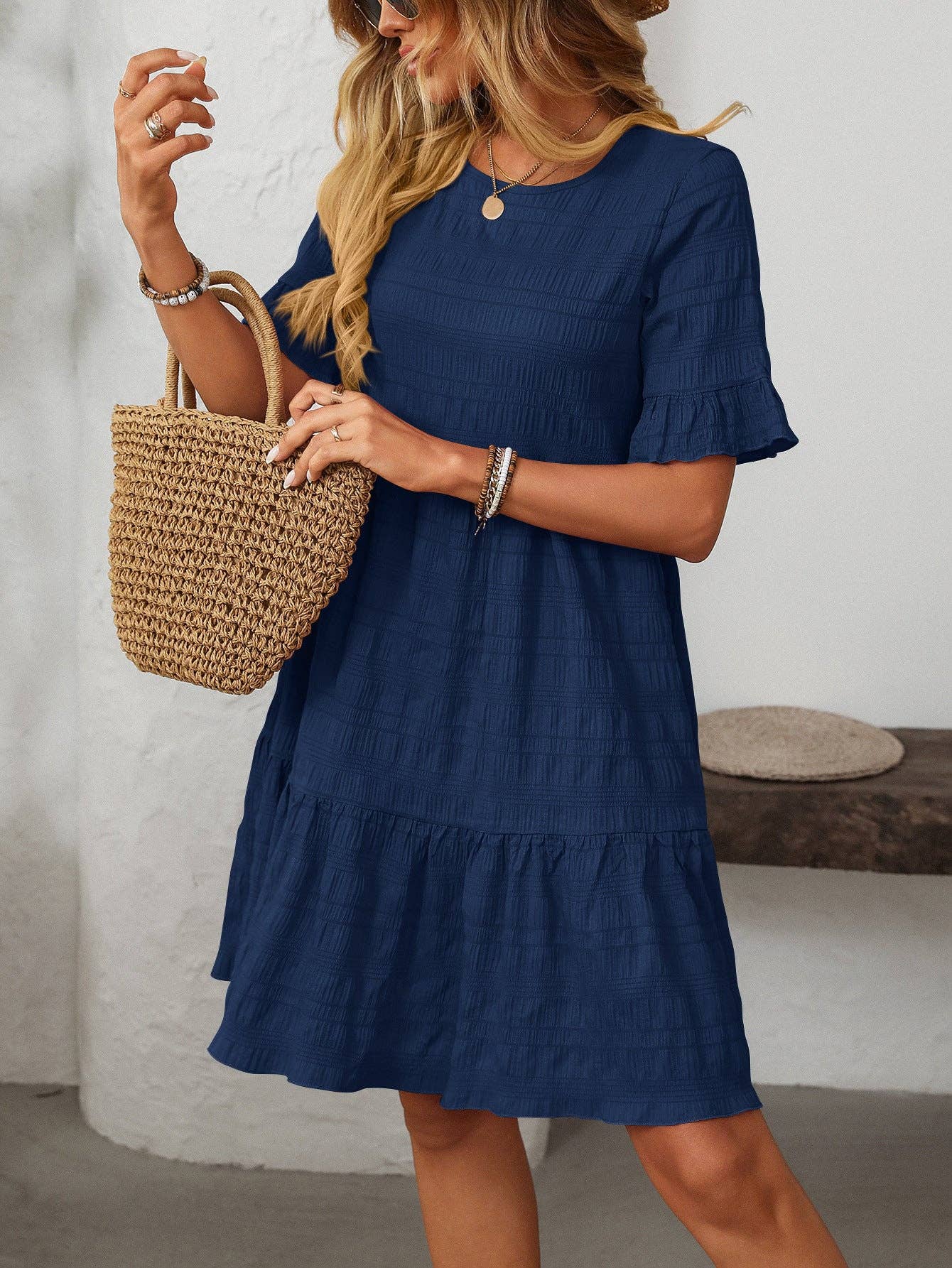 A woman stands indoors by a white textured wall and wooden bench, wearing Suhkasana’s Loose Casual Short-Sleeve Dress in navy blue and holding a woven straw bag—an effortlessly chic summer look.
