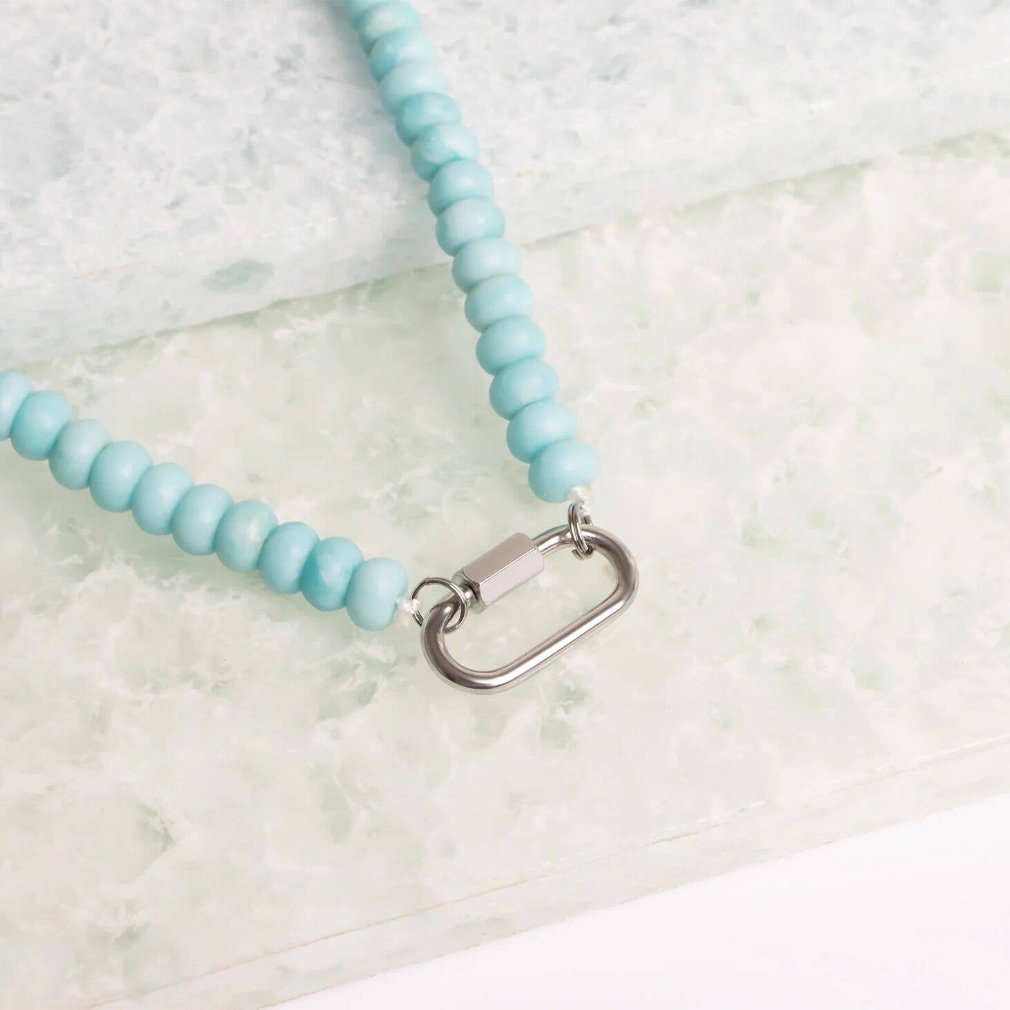 Close-up of the WJW Stainless Steel Turquoise Chunky Bead Carabiner Necklace, featuring light blue beads and a silver rectangular carabiner clasp, displayed on a textured white and pale green surface.