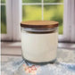 A 20oz, 3-wick soy wax candle by Stephanie Henry Candle Company, with a wood lid and coconut lime scent, rests on floral fabric in front of a window showcasing greenery, exuding calmness.