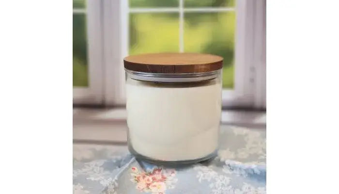 A 20oz, 3-wick soy wax candle by Stephanie Henry Candle Company, with a wood lid and coconut lime scent, rests on floral fabric in front of a window showcasing greenery, exuding calmness.