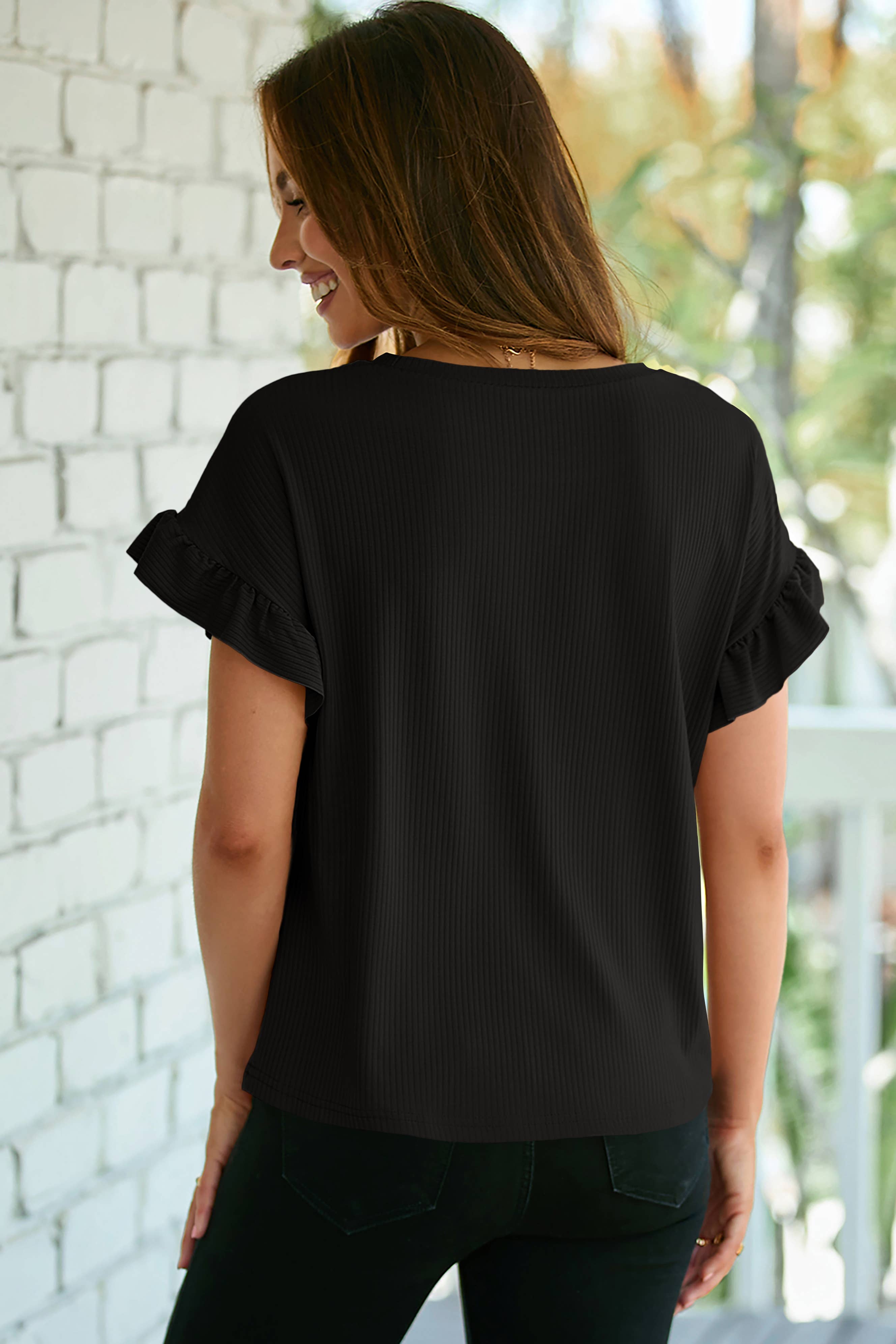 A woman stands on a porch, facing away from the camera, wearing the 1 the Woman Solid Color Ribbed Ruffled Short Sleeve T Shirt and black pants. A white brick wall and outdoor greenery create a stylish, feminine backdrop.