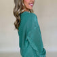 A woman with long hair smiles, wearing the Shewin Notched Neck Exposed Seam Drop Shoulder Sweatshirt with a comfortable green patchwork design and white pants, standing against a plain background.