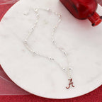 Seasons Jewelry's Alabama Glitter Enamel Logo Necklace, featuring a silver chain with round beads and a crimson “A” logo pendant in glitter enamel, is displayed on a round white marble surface beside a red glass bottle.