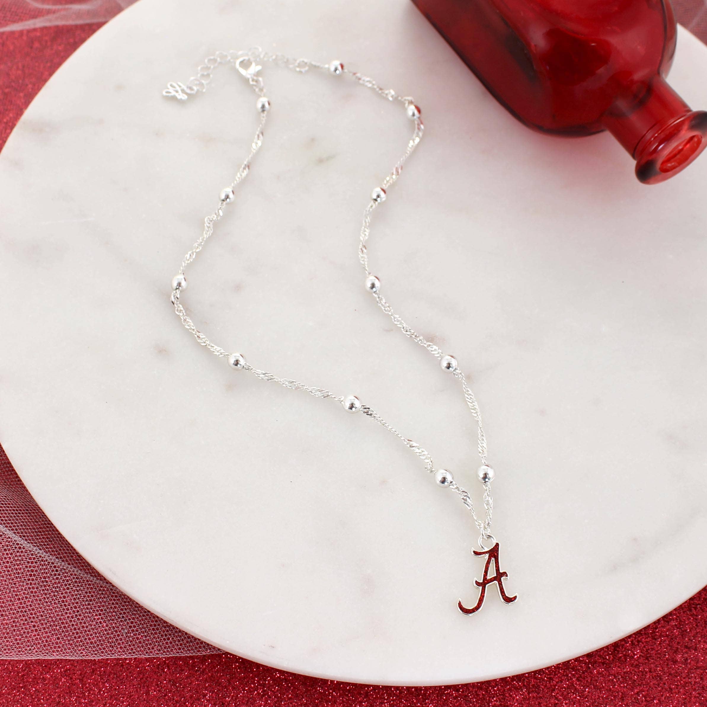 Seasons Jewelry's Alabama Glitter Enamel Logo Necklace, featuring a silver chain with round beads and a crimson “A” logo pendant in glitter enamel, is displayed on a round white marble surface beside a red glass bottle.