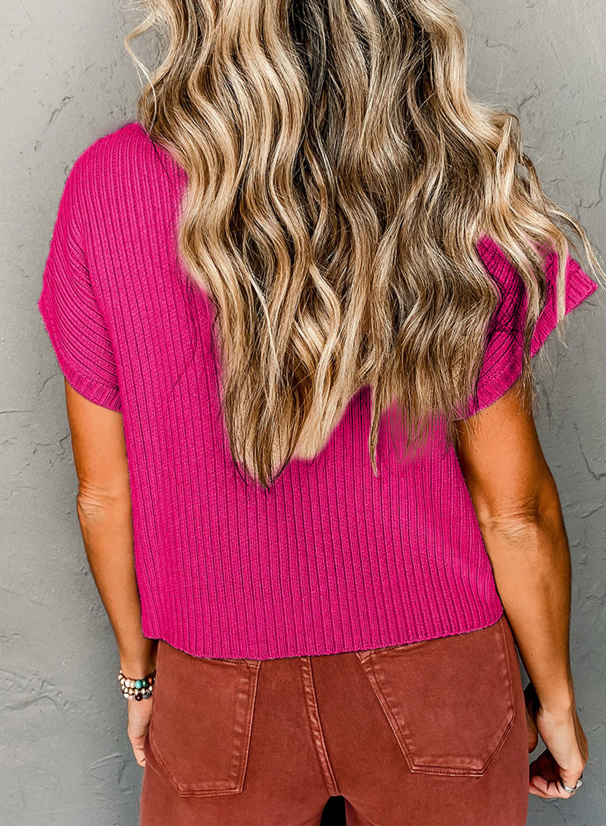 Someone with long wavy hair wearing a Shewin Patch Pocket Ribbed Knit Short Sleeve Sweater in pink and brown pants stands facing a gray wall, perfectly dressed for transitional weather.