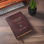 A brown, faux leather Devotional Every Day with Jesus book lying on a wooden desk next to a keyboard, pen, and small potted succulent.