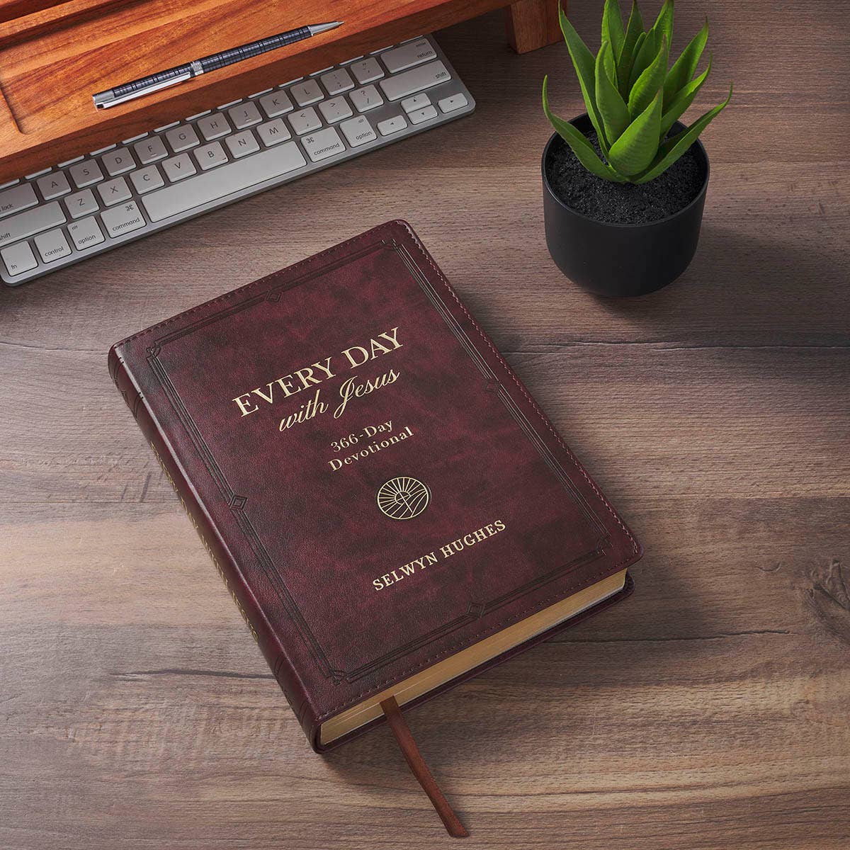 A brown, faux leather Devotional Every Day with Jesus book lying on a wooden desk next to a keyboard, pen, and small potted succulent.