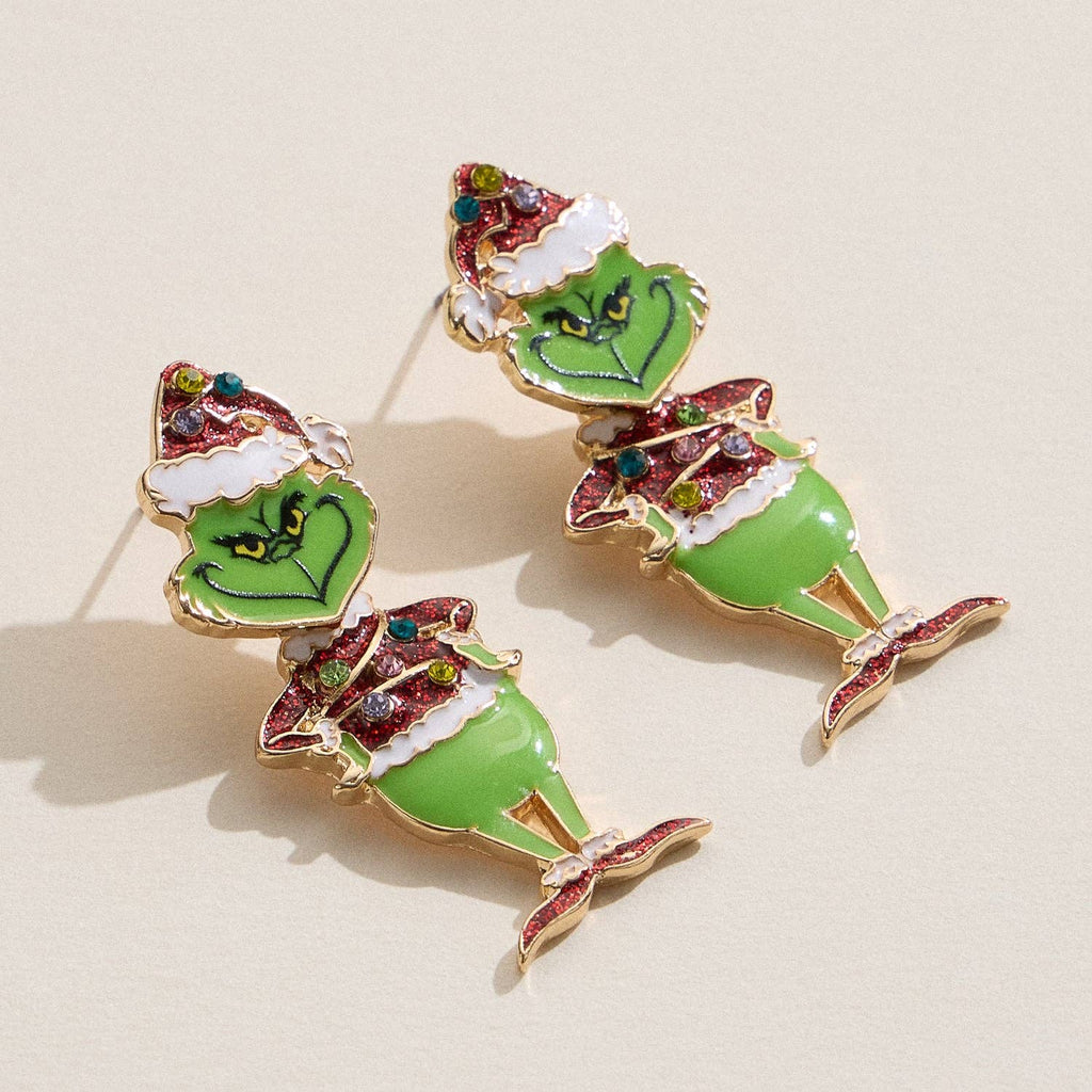 Add a festive touch to your look with the Enamel Green Monster Drop Earrings from Lilla Haven—these glitter holiday character earrings feature the mischievous figure in a Santa hat, detailed with green and red enamel, and are shown lying on a light background for the perfect seasonal accessory.