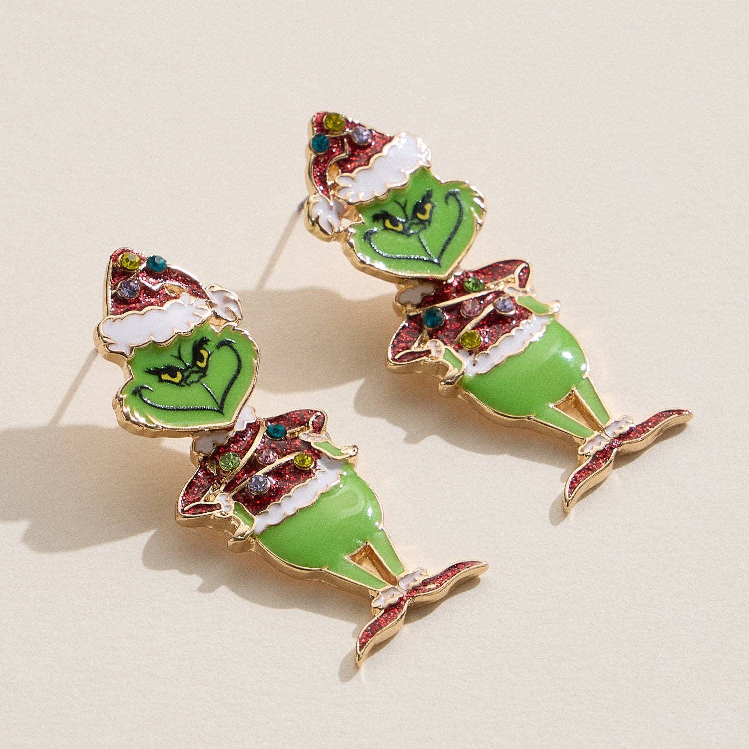 Add a festive touch to your look with the Enamel Green Monster Drop Earrings from Lilla Haven—these glitter holiday character earrings feature the mischievous figure in a Santa hat, detailed with green and red enamel, and are shown lying on a light background for the perfect seasonal accessory.