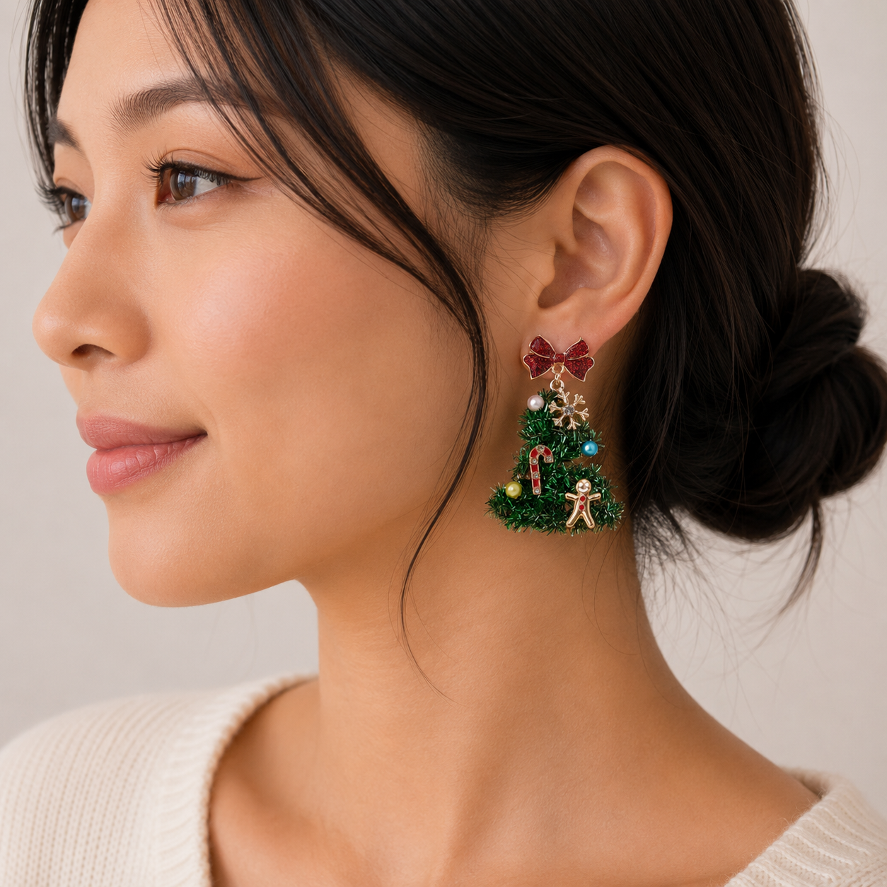 A woman with dark hair in a low bun wears Lilla Haven’s Tinsel Christmas Tree Drop Earrings, featuring holiday ornament charms, a red bow, and a gingerbread man. Styled with a light-colored sweater, her look is the perfect choice for anyone who loves playful festive jewelry.