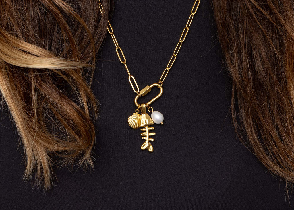 WJW's 18K Gold PVD Paperclip Carabiner Necklace features fishbone, shell, and pearl-style charms reminiscent of a customizable necklace. This gold chain piece highlights the on-trend paperclip design and is shown styled over a black top with brown hair partially visible.