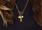 WJW's 18K Gold PVD Paperclip Carabiner Necklace features fishbone, shell, and pearl-style charms reminiscent of a customizable necklace. This gold chain piece highlights the on-trend paperclip design and is shown styled over a black top with brown hair partially visible.