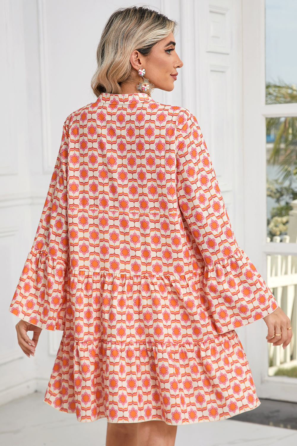 A woman is wearing the Shewin Orange Floral Button V Neck Bell Sleeve Mini Dress in a bright room with large windows.