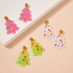 Six Christmas tree-shaped ornaments in pink, white, and green with colorful star decorations and gold star toppers are arranged on a beige surface and a white platform—reminiscent of Lilla Haven’s Glittery Christmas Tree Drop Earrings, festive holiday acrylic earrings designed to add sparkling cheer to your look.