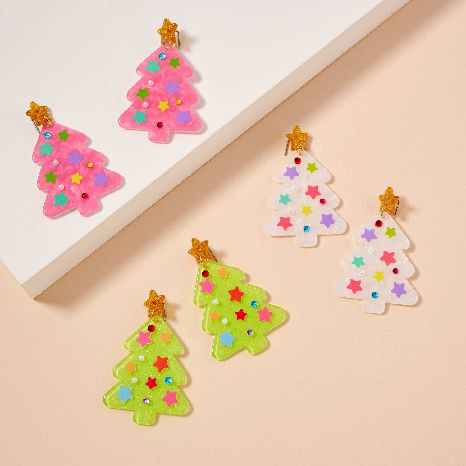 Six Christmas tree-shaped ornaments in pink, white, and green with colorful star decorations and gold star toppers are arranged on a beige surface and a white platform—reminiscent of Lilla Haven’s Glittery Christmas Tree Drop Earrings, festive holiday acrylic earrings designed to add sparkling cheer to your look.