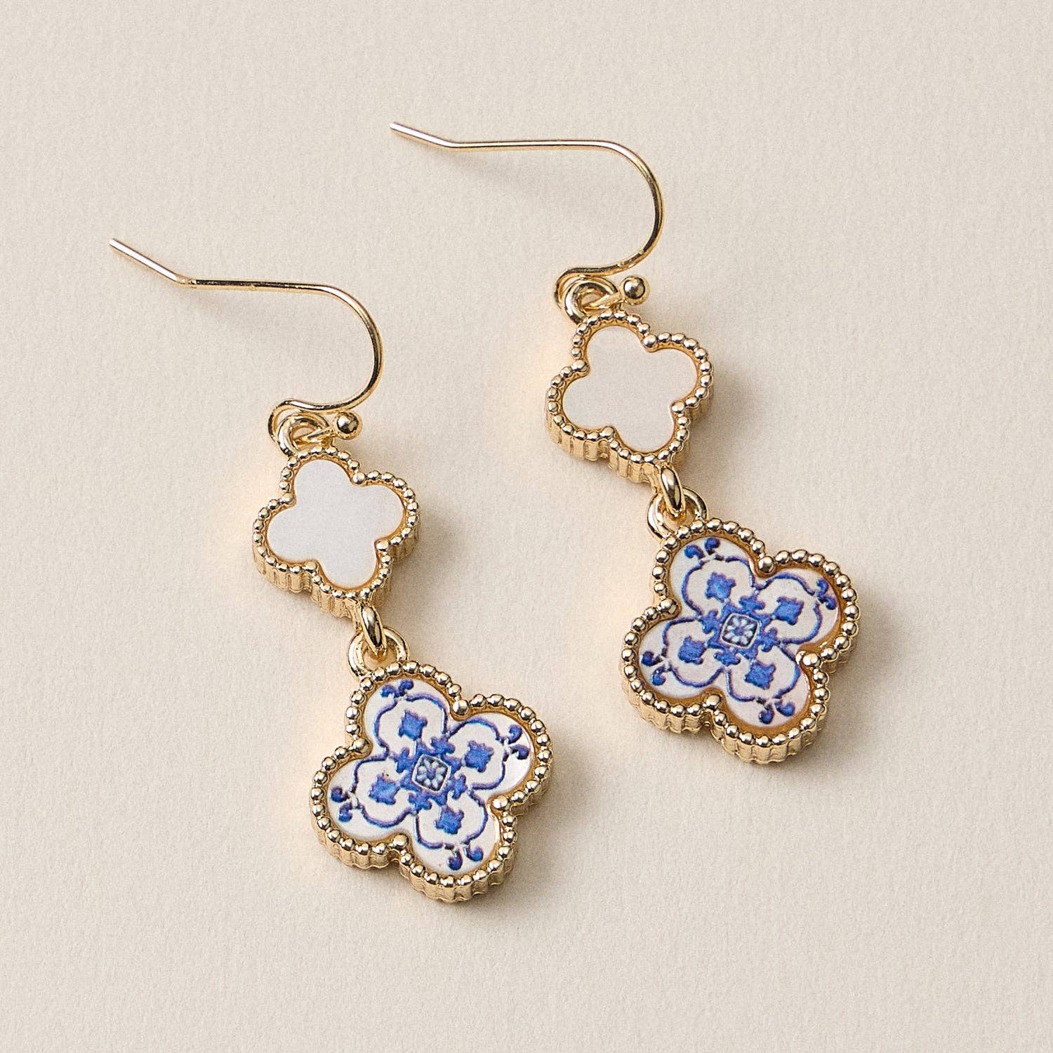 Lilla Haven’s Blue Enamel Clover Drop Earrings feature a graceful quatrefoil shape adorned with a blue floral motif on a white enamel background, finished with elegant gold trim and shown resting on a neutral surface. Main image