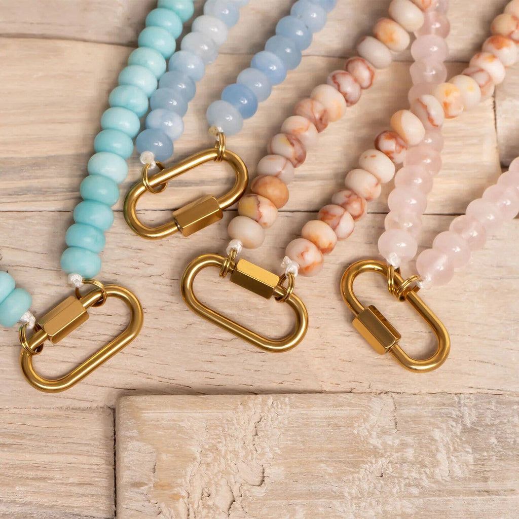 Four beaded lanyards in pastel blue, light blue, cream, and pink with gold clasps are arranged side by side on a wooden surface, their soft tones evoking the charm of the WJW Stainless Steel Turquoise Chunky Bead Carabiner Necklace.