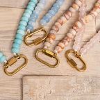 Four beaded lanyards in pastel blue, light blue, cream, and pink with gold clasps are arranged side by side on a wooden surface, their soft tones evoking the charm of the WJW Stainless Steel Turquoise Chunky Bead Carabiner Necklace.