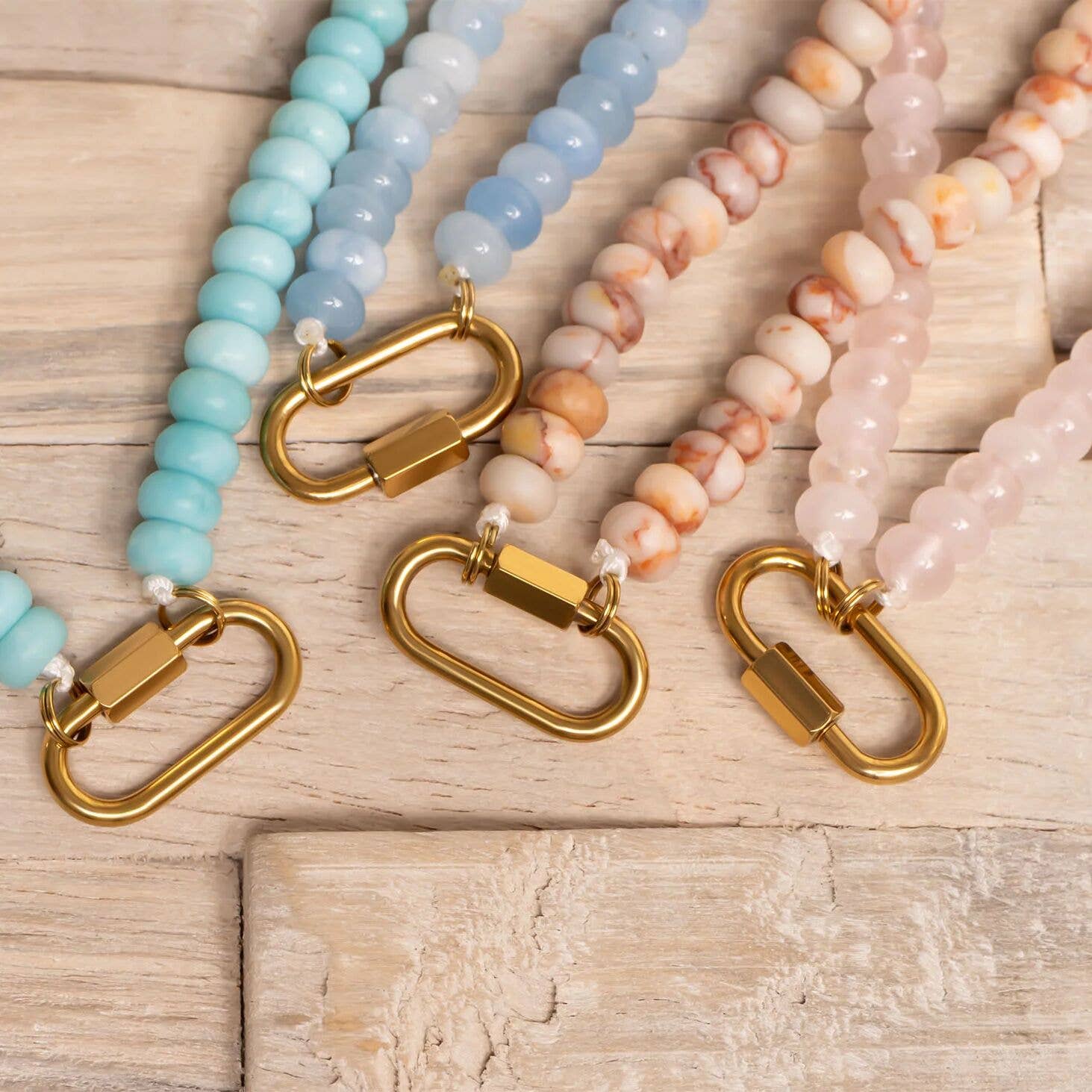Four beaded lanyards in pastel blue, light blue, cream, and pink with gold clasps are arranged side by side on a wooden surface, their soft tones evoking the charm of the WJW Stainless Steel Turquoise Chunky Bead Carabiner Necklace.