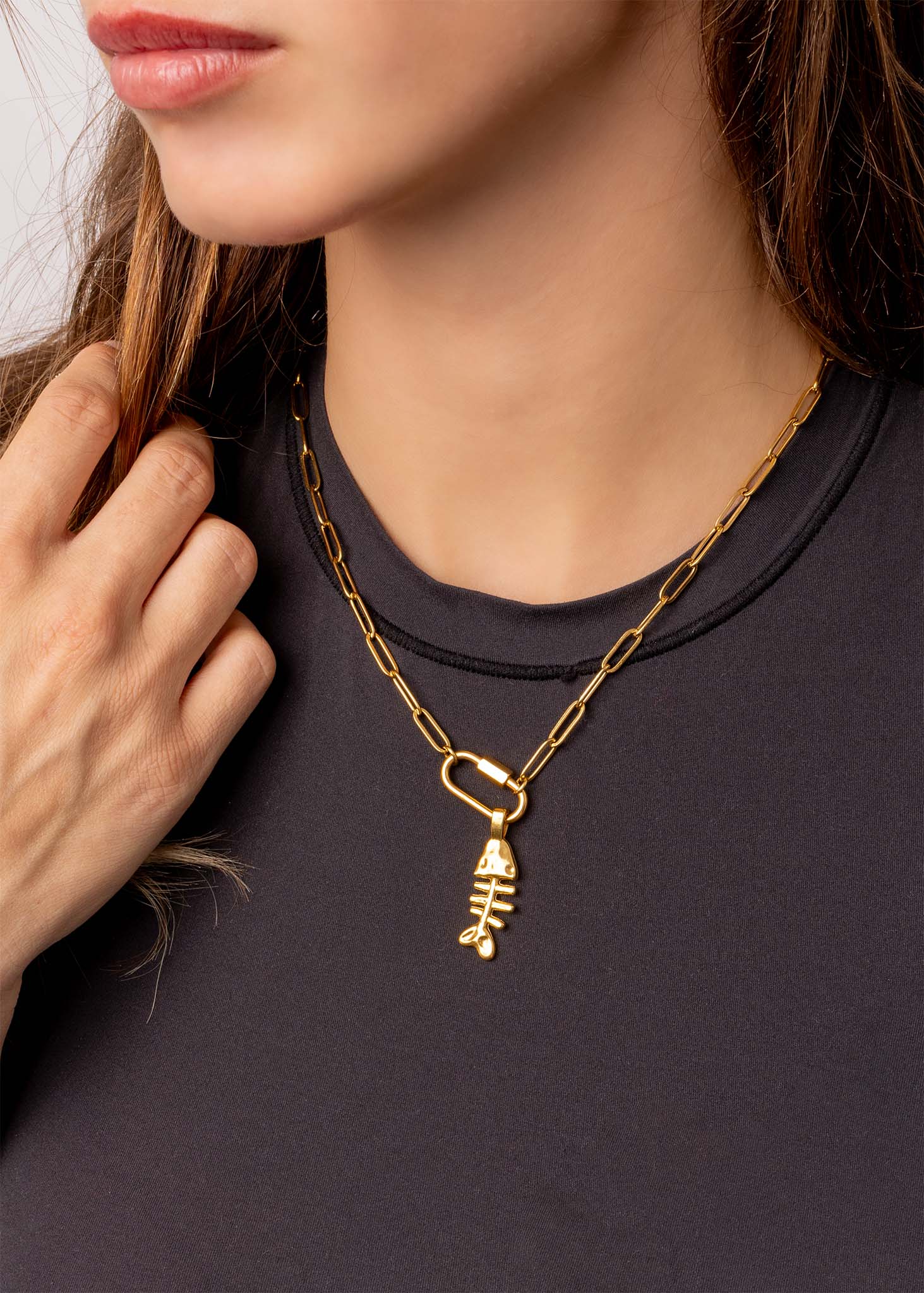 A woman wearing a black top and a WJW 18K Gold PVD Paperclip Carabiner Necklace with a unique geometric pendant holds the customizable necklace in her left hand.