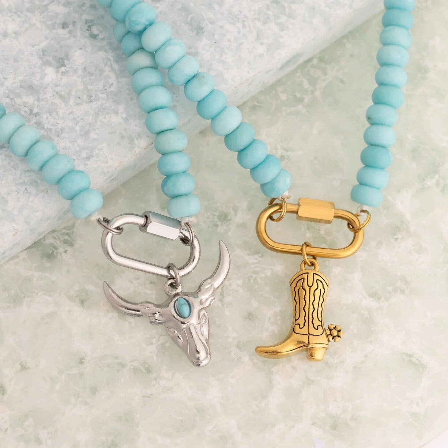 Two WJW Stainless Steel Turquoise Chunky Bead Carabiner Necklaces are displayed on a light stone surface—one featuring a silver bull skull pendant, and the other adorned with a gold cowboy boot pendant. Each unique necklace is crafted with durable stainless steel components for lasting wear.