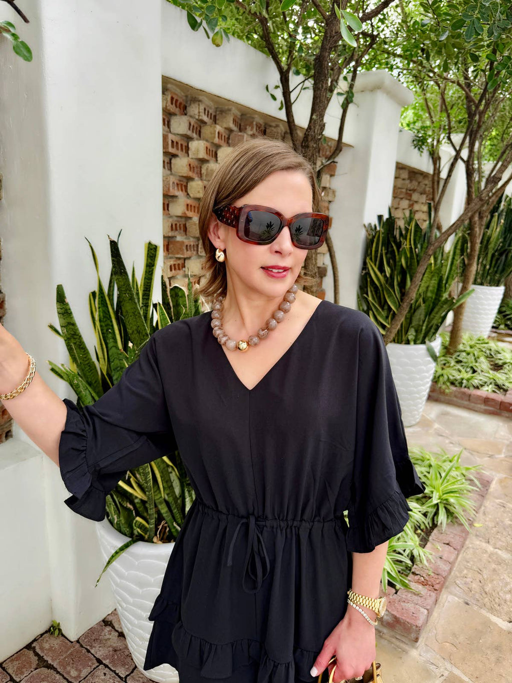 A woman wearing a black dress, large sunglasses, and the Marjorie Fall Beaded Necklace by Canvas Style stands outdoors near potted plants and a white wall.
