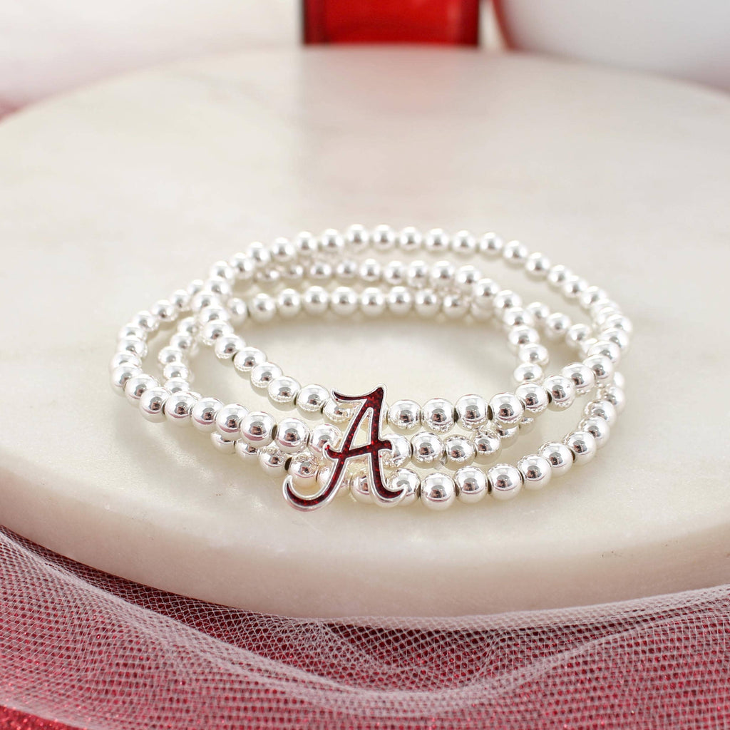 The Alabama Glitter Enamel Logo Stretch Bracelet Set by Seasons Jewelry, featuring silver beads and a red and silver “A” charm accented with crimson glitter, is displayed on a white round surface.
