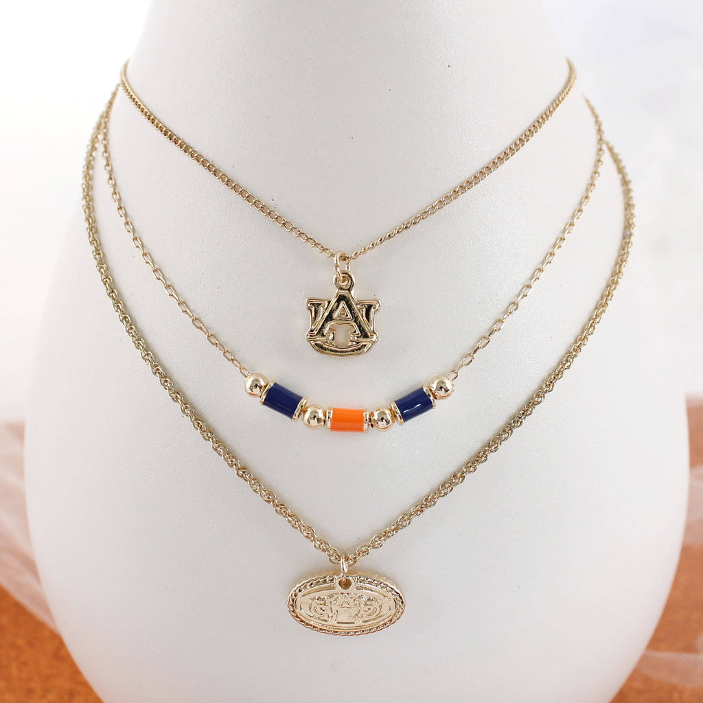 Discover the Auburn Gold & Tube Bead Trio Necklace Set by Seasons Jewelry: a stunning three-layered necklace displayed on a stand. The top layer features an "A" pendant, the middle showcases orange and blue tube beads inspired by Auburn University designs, and the bottom displays an oval medallion with an intricate design—a perfect War Eagle jewelry statement piece.