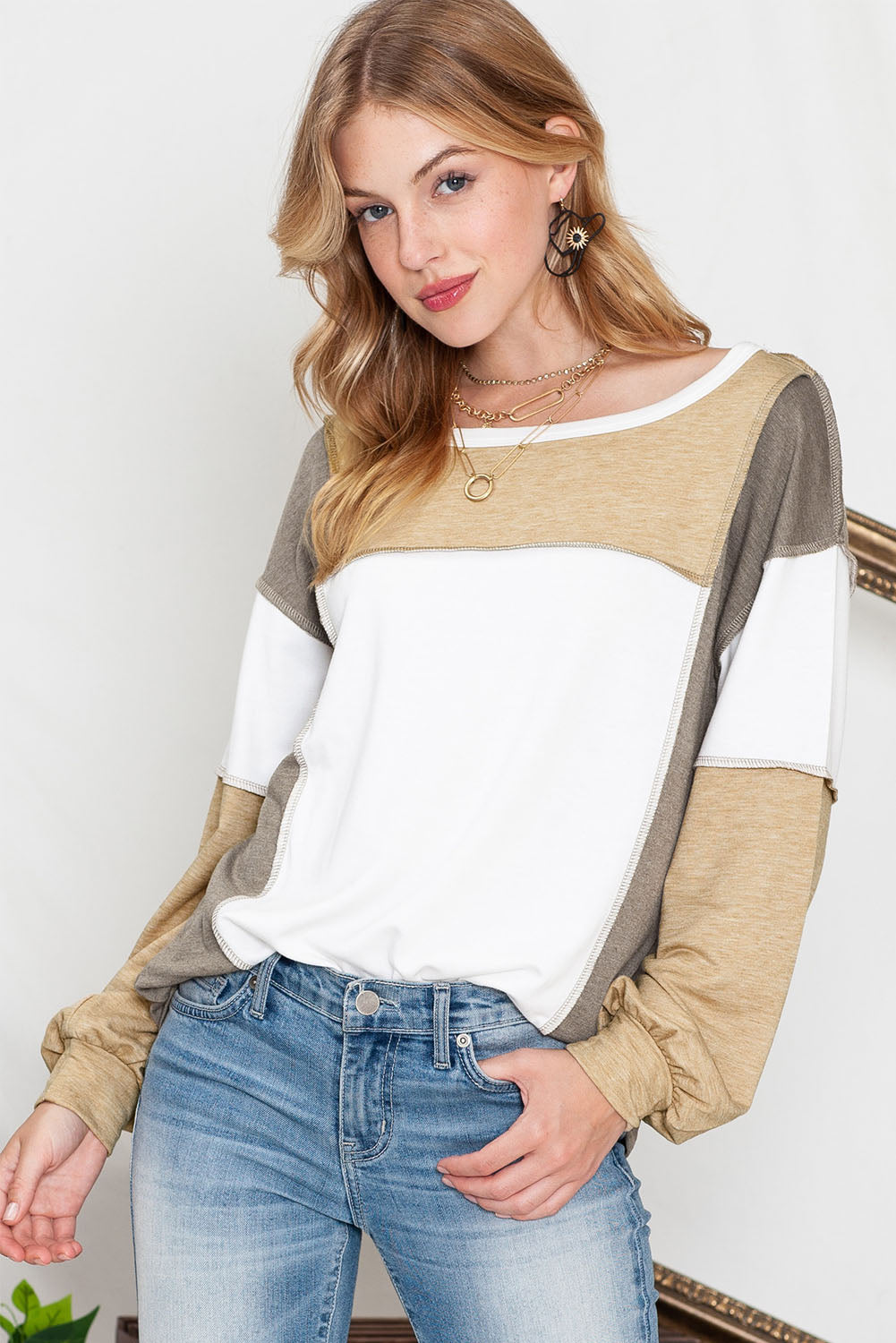 A person wearing the Shewin Khaki Color Block Exposed Seam Long Sleeve Top paired with jeans, featuring a distinctive patchwork design, accessorized with layered necklaces and earrings, stands against a neutral background.