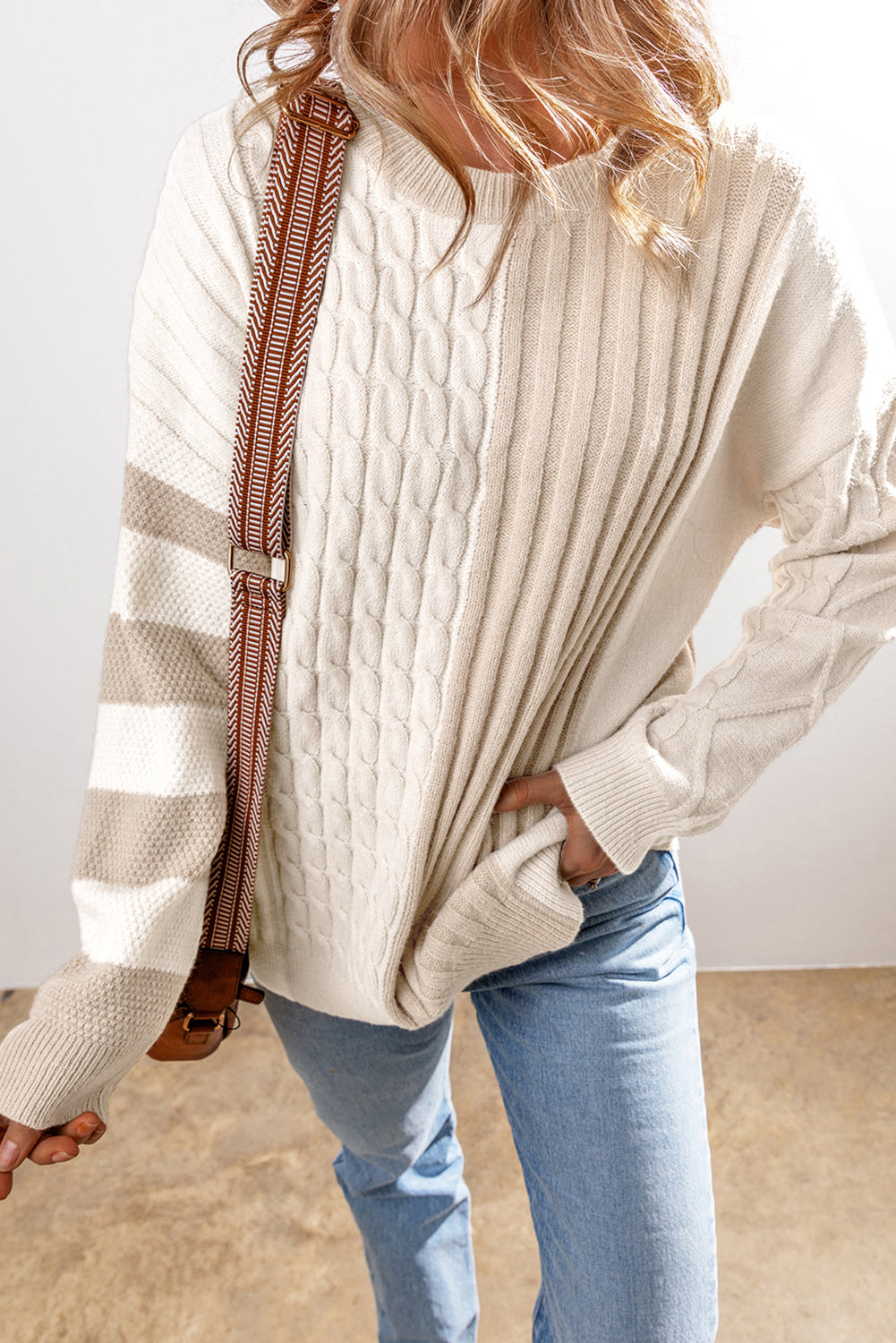 A person wearing a Shewin Khaki Cable Knit Colorblock Crew Neck Drop Shoulder Sweater with blue jeans stands with a crossbody bag.