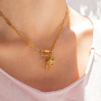 A person wearing the WJW 18K Gold PVD Paperclip Carabiner Necklace with three charms—a fish, a lock, and a feather—over a light pink shirt.