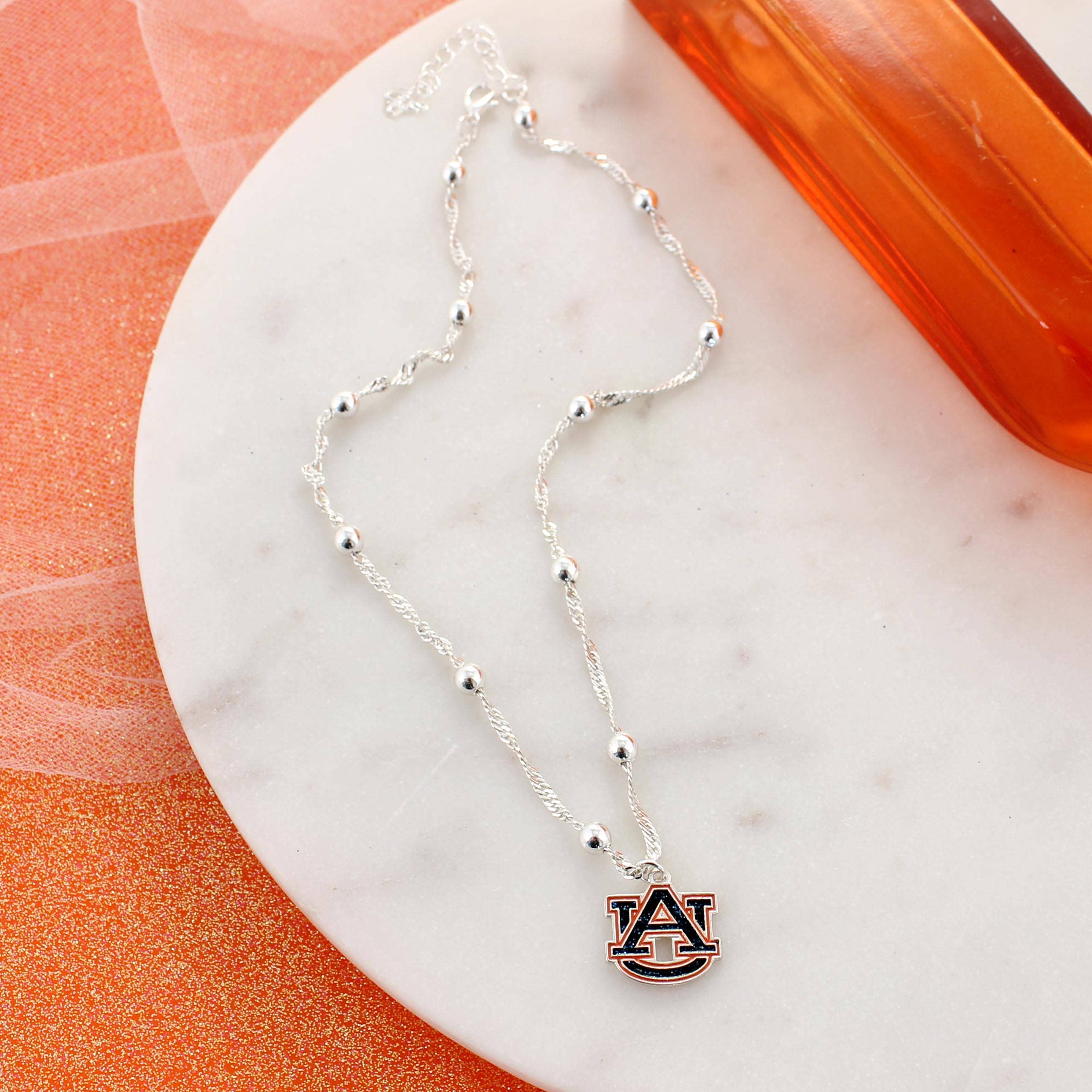 Auburn Glitter Enamel Logo Necklace by Seasons Jewelry, featuring a silver chain with small round beads and a glitter enamel AU logo pendant, displayed on a white marble surface alongside an orange container.