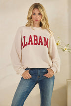 A woman wears an off-white, long-sleeve, textured knit ALABAMA Cloudy Soft Knit Long Sleeve Top with red athletic-style letters across the chest, paired with blue jeans.