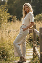A woman stands on a wooden path in a grassy setting, wearing a white short-sleeve top, Sewn and Seen's Woven Striped Wide Leg Pants, and sandals.