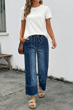 A woman stands outdoors wearing 1 the Woman Drawstring Seam Detail Raw Hem Wide Leg Denim Pants with an adjustable waist, paired with a white top, sandals, and a brown crossbody bag.