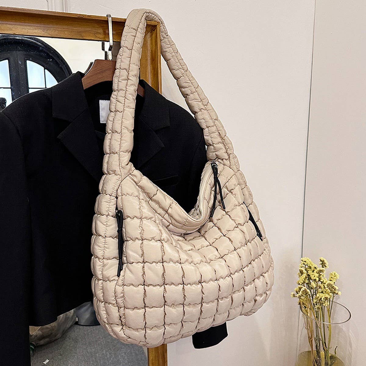 A cream-colored ACCITY Cloud Tote Bag Large Shoulder Crossbody with quilted design and zipper hangs on a black blazer by a wooden mirror. A small vase of yellow flowers nearby adds charm to this trendy display.