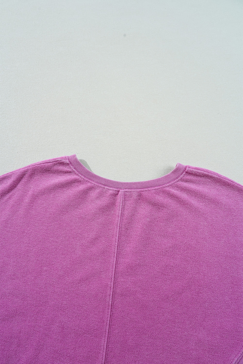 A close-up of the Shewin Notched Neck Exposed Seam Drop Shoulder Sweatshirt in pink, laid flat on a light surface, highlights its back and sleeve details in high-quality fabric.