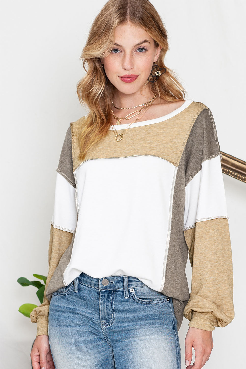 A woman with long hair is wearing the Shewin Khaki Color Block Exposed Seam Long Sleeve Top, featuring beige, white, and olive panels in a unique patchwork design. She stands against an indoor backdrop with green leaves partially visible to the side, paired perfectly with light blue jeans.