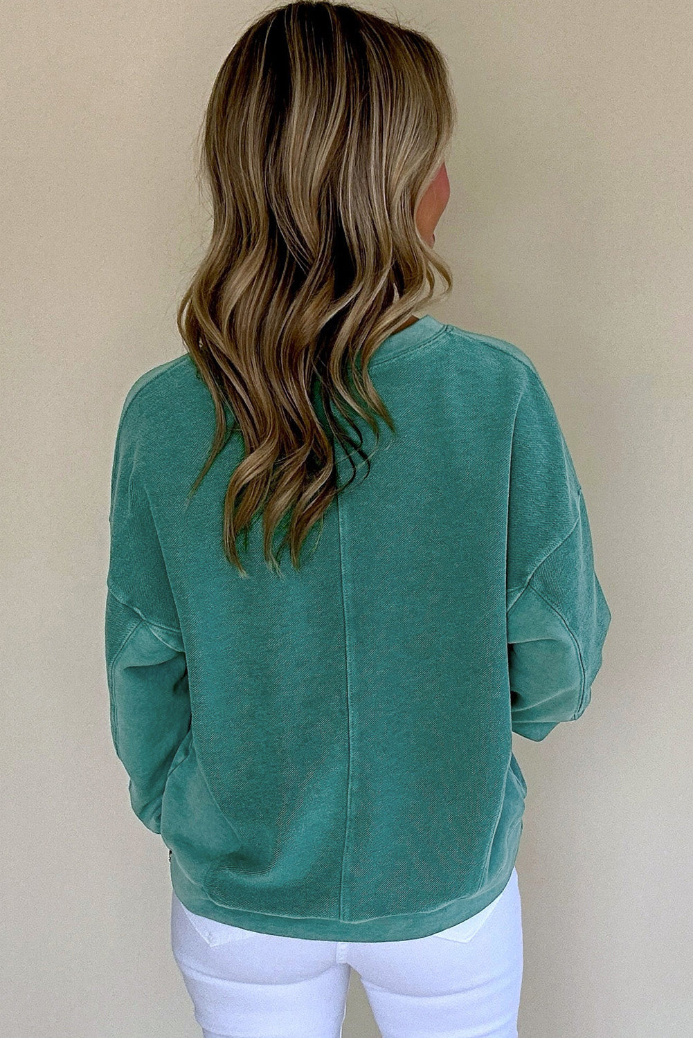 A person with long, wavy hair wears a Shewin Notched Neck Exposed Seam Drop Shoulder Sweatshirt against a plain background, paired with white pants.