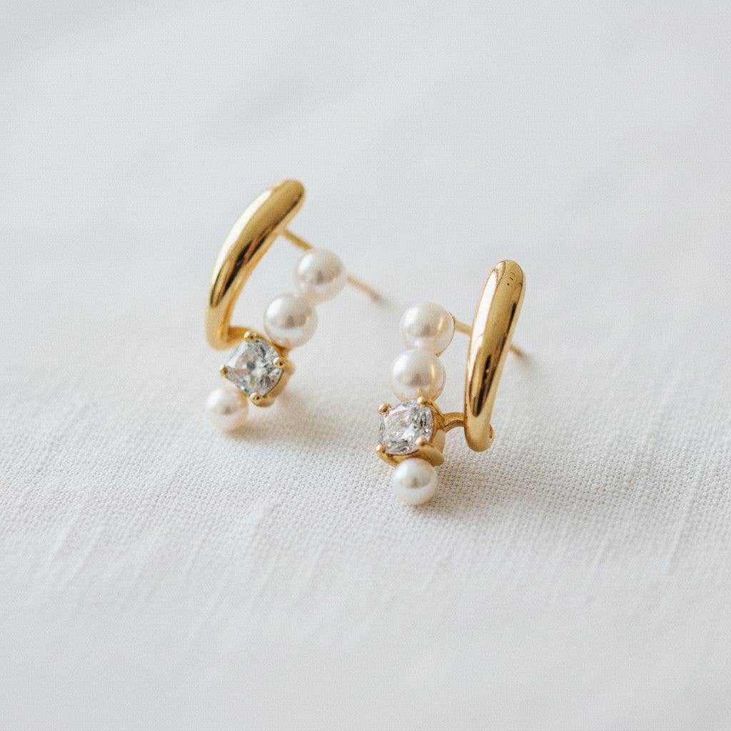 The Ellemadera Zircon Pearl Dangle Earrings feature a gold design adorned with small white pearls and sparkling clear gemstones, elegantly displayed on a white fabric background—an exquisite example of refined jewelry.