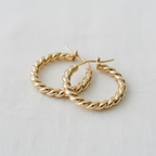 The Ellemadera Sculptural Twisted Hoop Earrings, featuring twisted gold hoops and sleek silver posts, are displayed on a white fabric background, embodying the elegance of minimalist earrings.