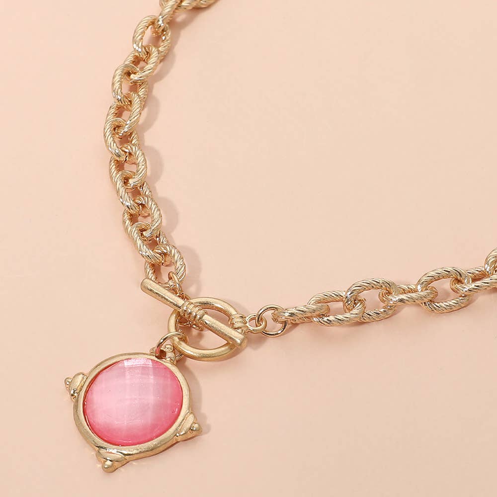 Sensibling Corp.’s Faceted Round Stone Charm Pendant Toggle Necklace, featuring a gold chain with a round pink pendant and toggle-style closure, displayed on a light beige background.