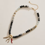 The Textured Glass Bead Necklace with Charm Trio by Lilla Haven features black, white, and clear glass beads accented with gold-toned spacers, and showcases three dangling charms—a wishbone, a red horn, and a gold shape—making this charm necklace an eye-catching accessory.