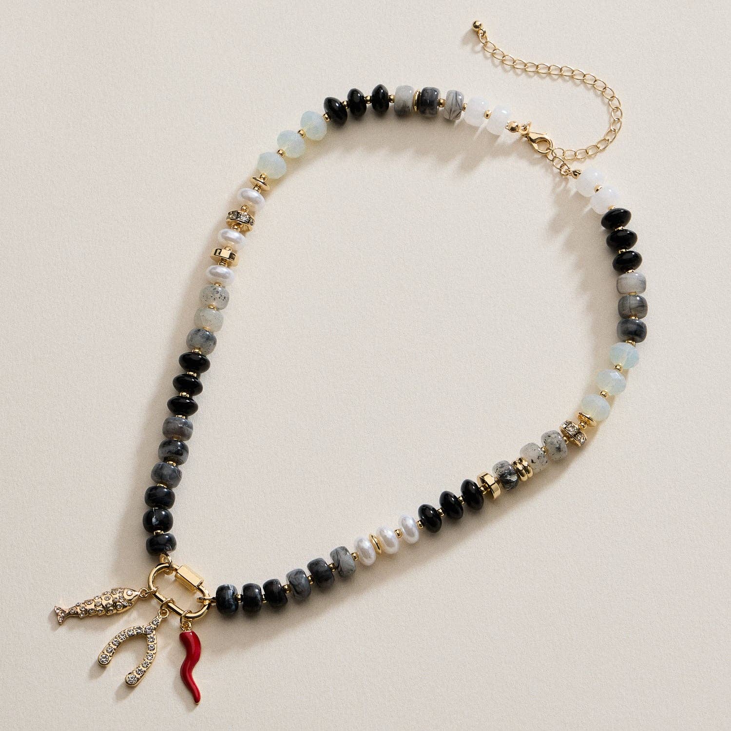 The Textured Glass Bead Necklace with Charm Trio by Lilla Haven features black, white, and clear glass beads accented with gold-toned spacers, and showcases three dangling charms—a wishbone, a red horn, and a gold shape—making this charm necklace an eye-catching accessory.