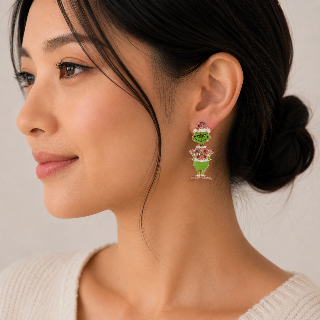 A woman in profile with her hair in a low bun is wearing a cream sweater and Lilla Haven’s Enamel Green Monster Drop Earrings—colorful, glittery holiday jewelry shaped like a green-skinned character with a pink hat holding a present, perfect for the festive season.