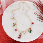 The Vintage Christmas Charm Necklace by Seasons Jewelry features gold-tone links adorned with festive red and gold holiday charms—a stocking, candy cane, ornament, and two brilliant red beads. Displayed on a round white marble surface with accents of red glitter and mesh, this necklace captures vintage-style elegance perfect for the Christmas season.