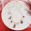 The Vintage Christmas Charm Necklace by Seasons Jewelry features gold-tone links adorned with festive red and gold holiday charms—a stocking, candy cane, ornament, and two brilliant red beads. Displayed on a round white marble surface with accents of red glitter and mesh, this necklace captures vintage-style elegance perfect for the Christmas season.