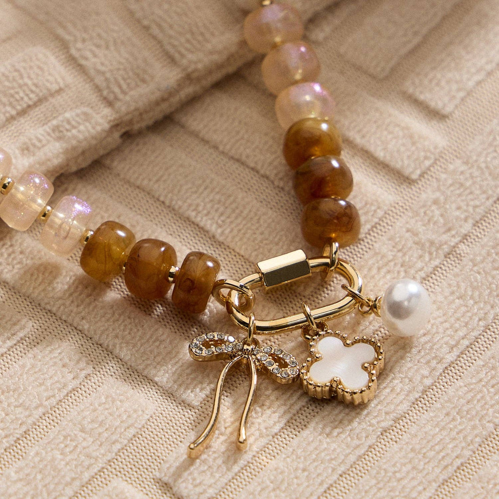 Textured Glass Bead Necklace with Charm Trio by Lilla Haven, featuring brown and cream glass beads accented with gold-toned spacers, and adorned with a trio of gold charms: a rhinestone-studded bow, a white flower, and a pearl—all elegantly displayed on textured beige fabric.